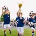 Happy boys in elementary school sports team celebrating soccer succes in tournament final game. Kids winning football game. Happy children sports team rising golden trophy